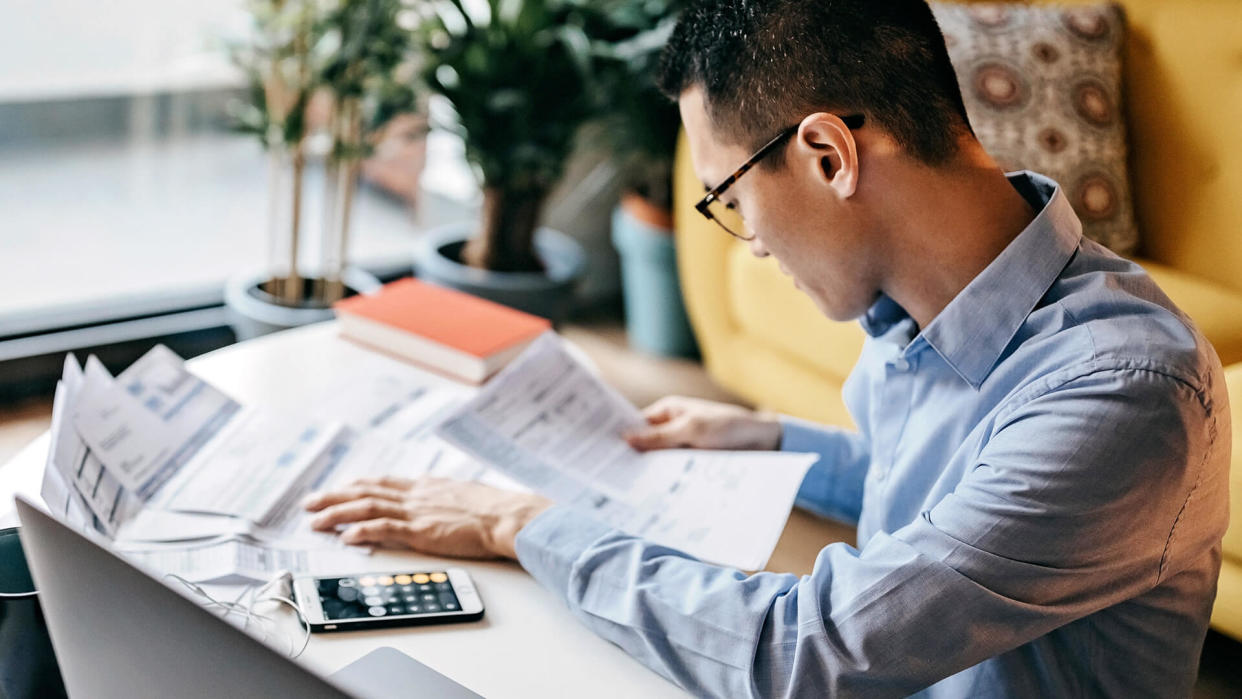 Japanese man with eyeglasses siting on floor in the living room and using smart phone and laptop for managing home finances.