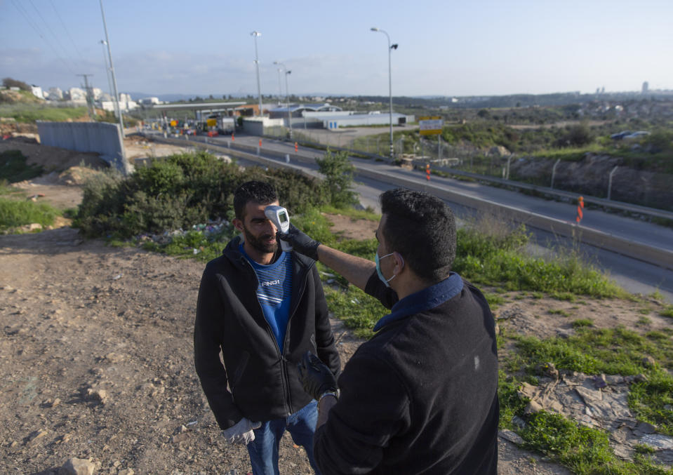 A paramedic from the Palestinian Ministry of Health checks the body temperature of a Palestinian laborer after exiting an Israeli army checkpoint, background, on his way home at the end of a day work in Israel, part of a strict precautionary measures to contain coronavirus outbreak, in the bordering West Bank village of Nilin, west of Ramallah, Wednesday, March 25, 2020. (AP Photo/Nasser Nasser)