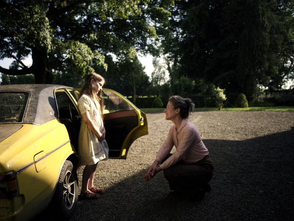 A woman crouches to talk to a young girl standing next to an open car door.