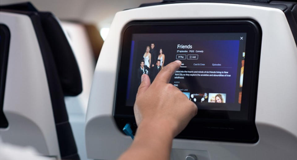 An inflight entertainment console is shown with a hand selecting to watch TV show 'Friends'.