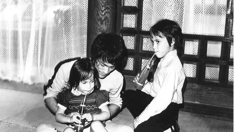 Bruce in yellow jumpsuit, cross legged with Shannon in his lap, Brandon kneeling to his left, children drinking soda. On set of GOD, pagoda.