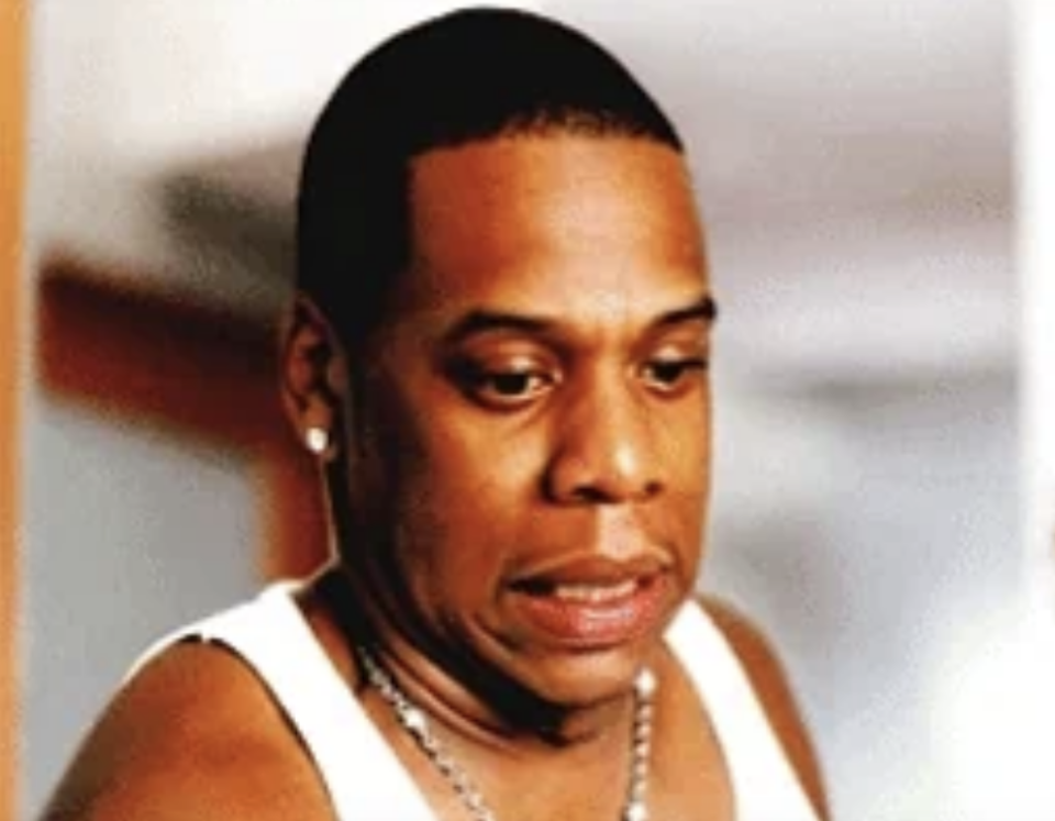 Jay-Z, wearing a sleeveless top and a necklace, appears pensive, looking down with a slightly furrowed brow in this candid image