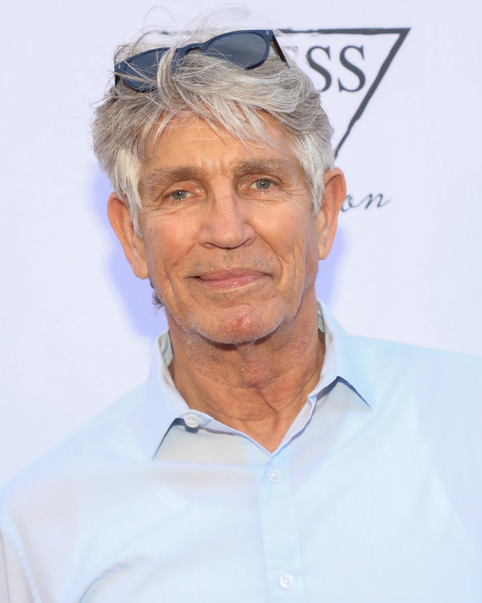 Eric Roberts smiles, wearing a light-colored shirt with sunglasses on his head, in front of a backdrop with partially visible text