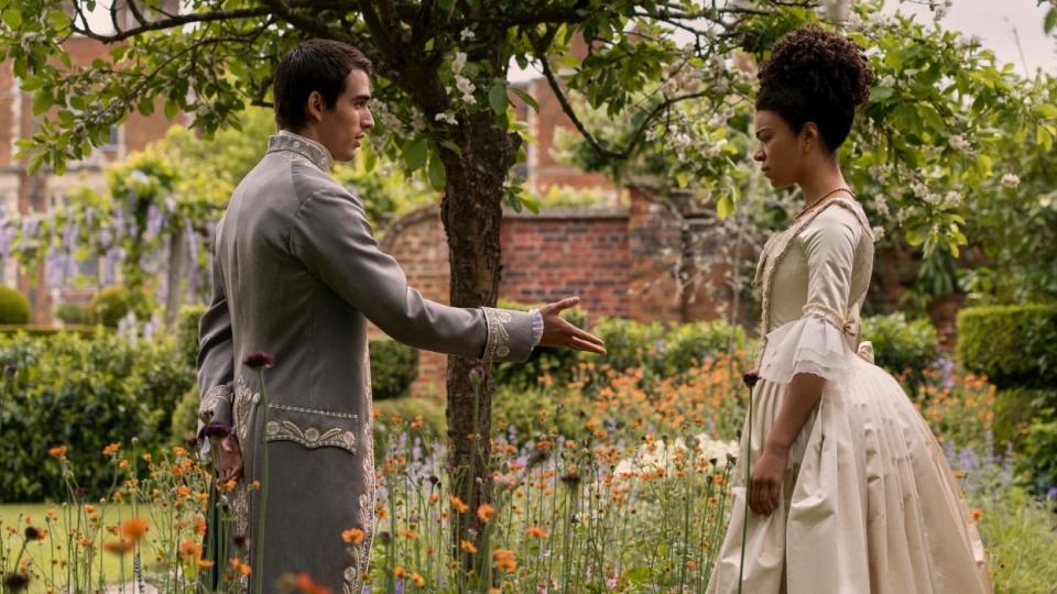 Man in period costume reaching out to woman in garden dress, both standing amidst flowers