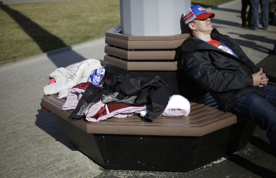 In this Wednesday, Feb. 12, 2014, photo, jackets sit on a bench as a spectator rests in the sun in the Olympic Park at the 2014 Winter Olympics, in Sochi, Russia. Temperatures were near 60 degrees Fahrenheit in Sochi on Wednesday. (AP Photo/David Goldman)