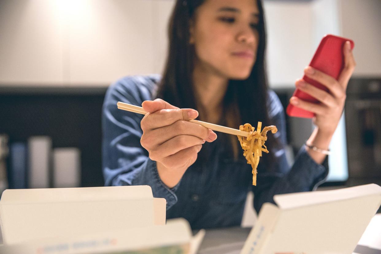 Selective focus of takeout chopsticks holding chow mein, woman looking at smartphone with a red case at a table with a wall in the background