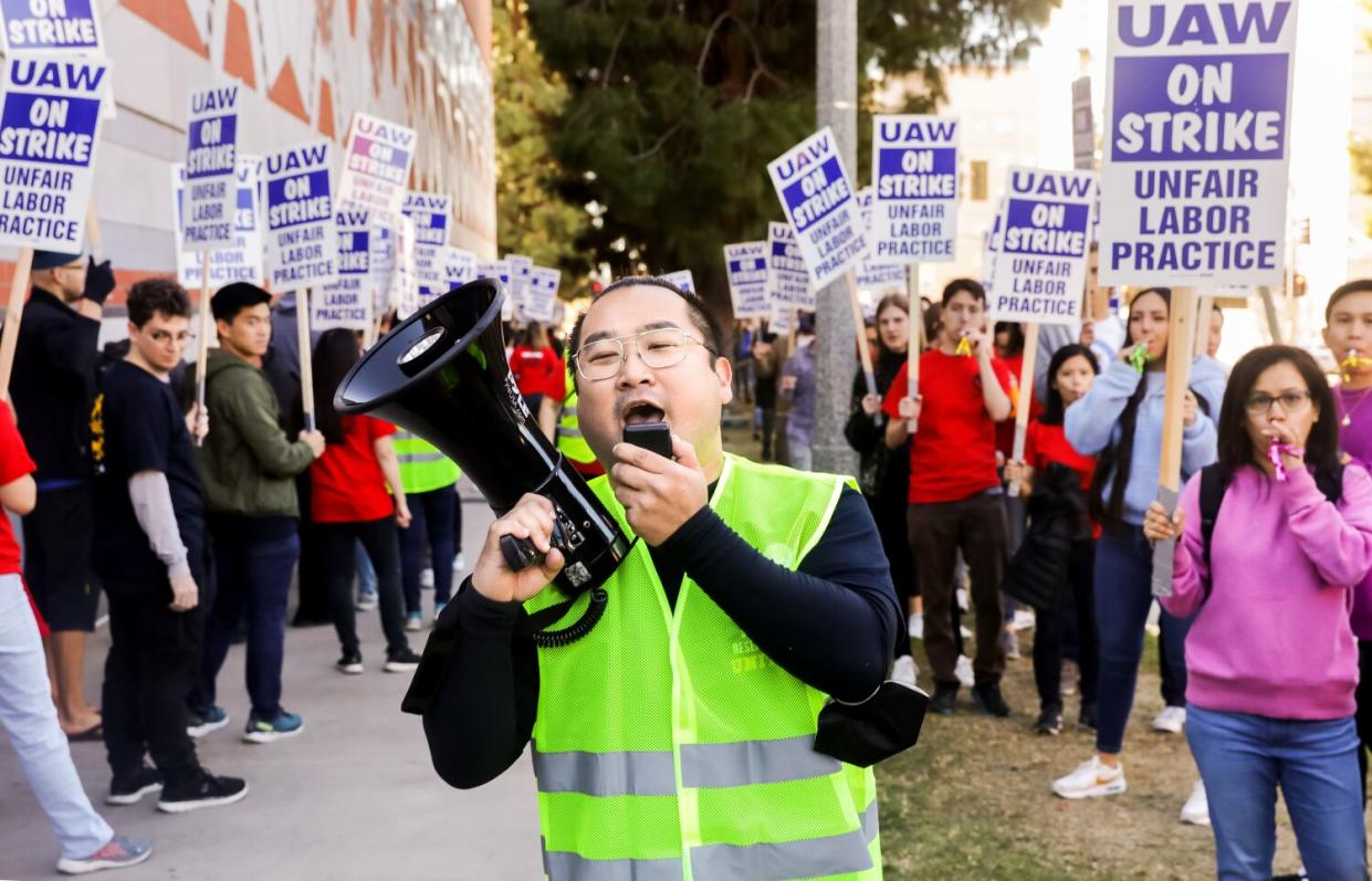 A man in a neon-colored safety vest talks into a megaphone as fellow demonstrators hold signs nearby.