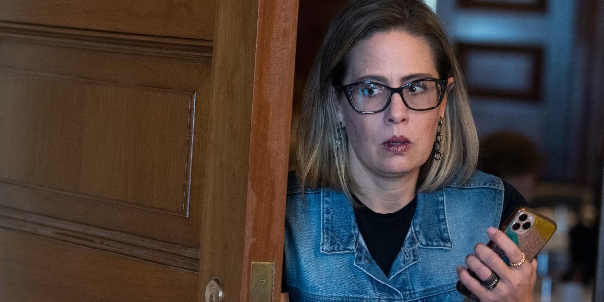 Kyrsten Sinema holds her phone up while exiting a wooden doorway wearing a jean jacket.