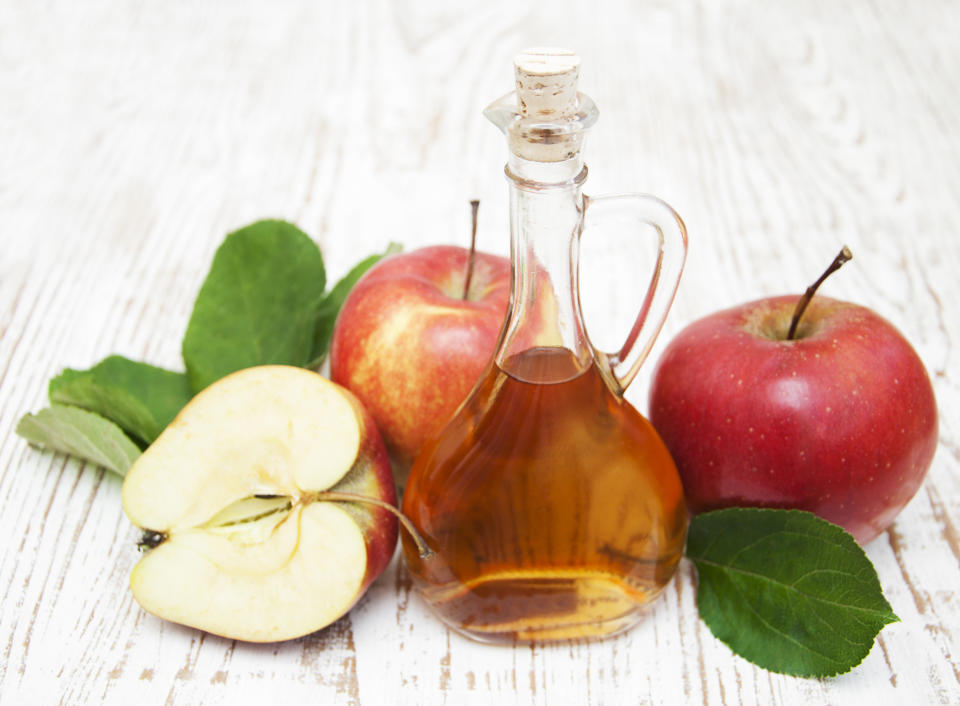 A bottle of apple cider vinegar on a white wooden table surrounded by apples