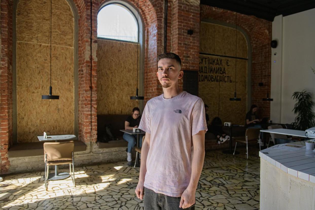 A bearded man in a beige T-shirt stands in a cafe with brick walls and boarded-up windows.