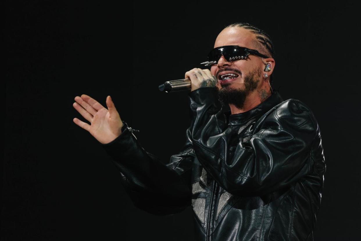 J Balvin wears a black leather jacket and shades and sings into a microphone