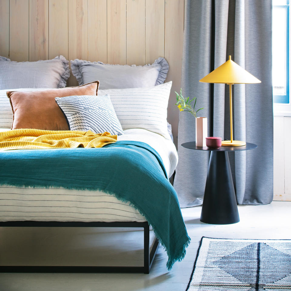  A bedroom with a bed dressed in multicoloured bedlinen and a bedside table holding a lamp with a cone-shpaed shade. 