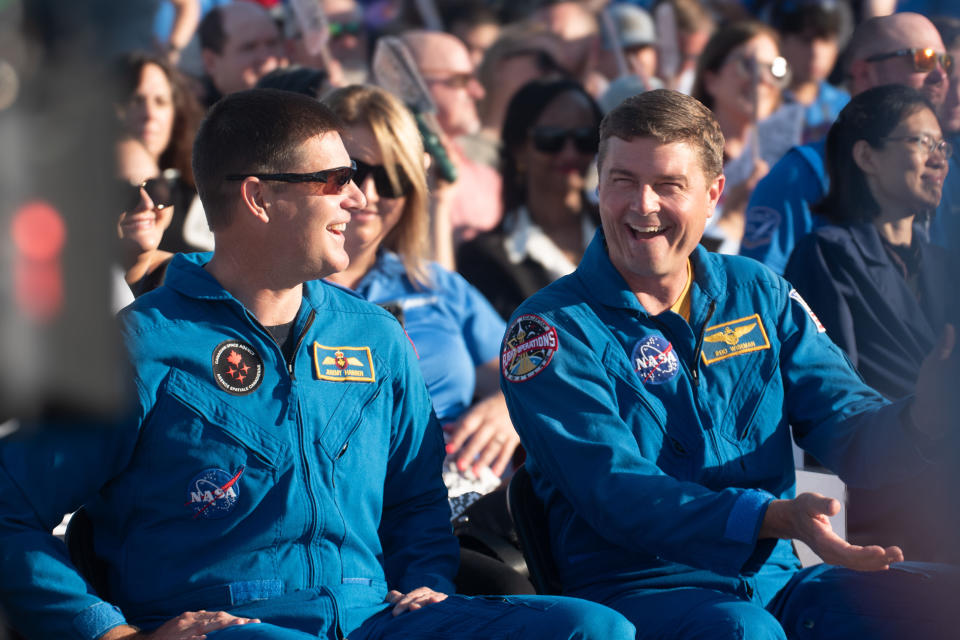 Zwei Männer in blauen Overalls lachen, während sie in einer Menschenmenge sitzen.