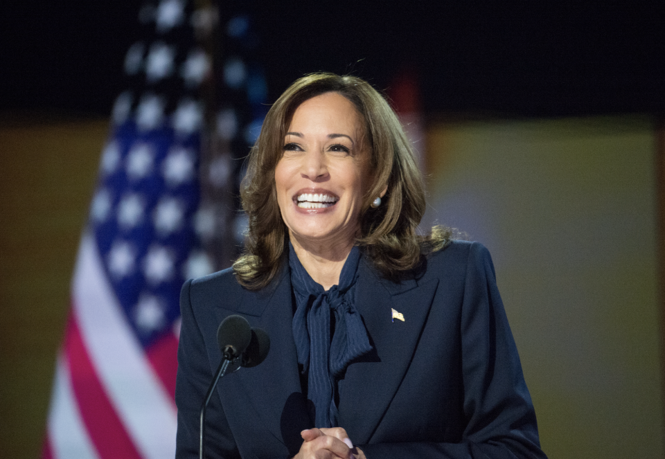 A smiling woman stands at a podium, wearing a professional suit, with an American flag in the background