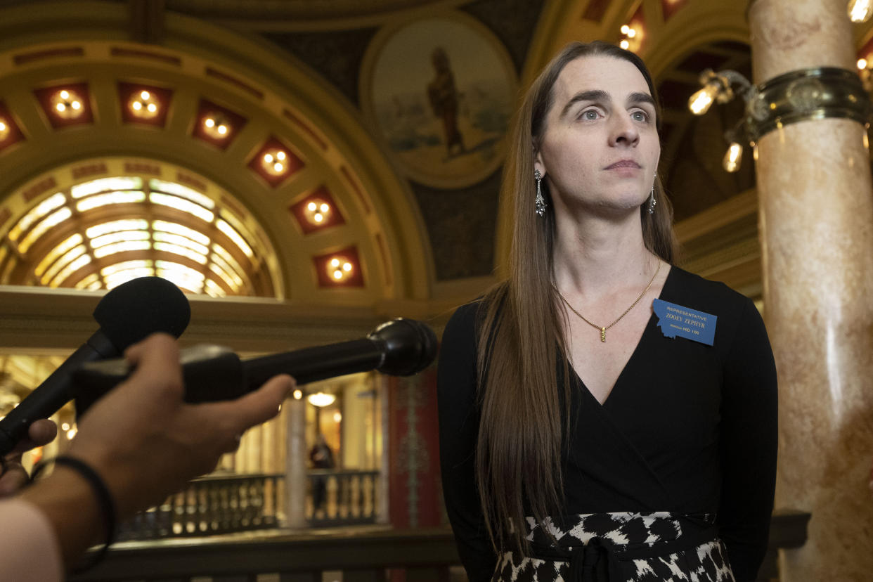 Rep. Zooey Zephyr stands in front of the outstretched microphones of reporters in a colonnaded hall with an arched gallery stretching behind.