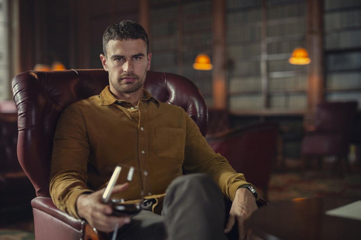 theo james in the gentlemen, a young man sits in an armchair looking at the camera and holding a wine glass