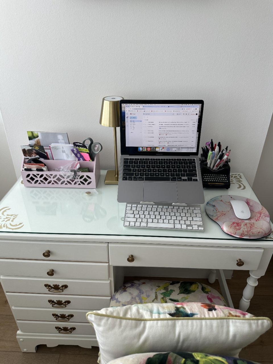 Home office setup with a white desk, laptop on a stand displaying emails, lamp, stationery holder, and a floral cushion on the chair