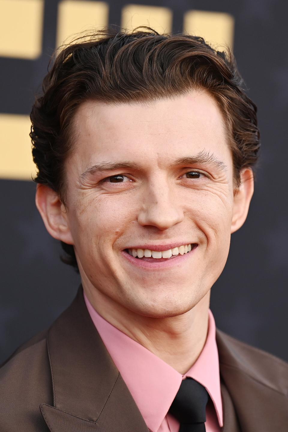 Tom Holland in a brown suit and pink shirt smiling at an event