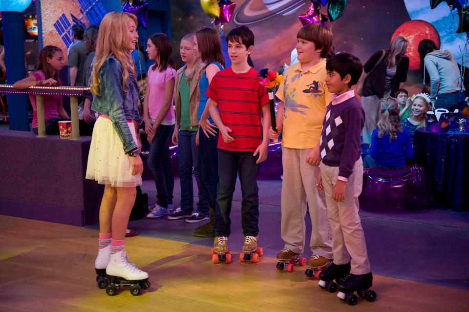 Girl in a white skirt and roller skates talking to three boys at a roller skating rink. People in the background are engaged in conversation and activities