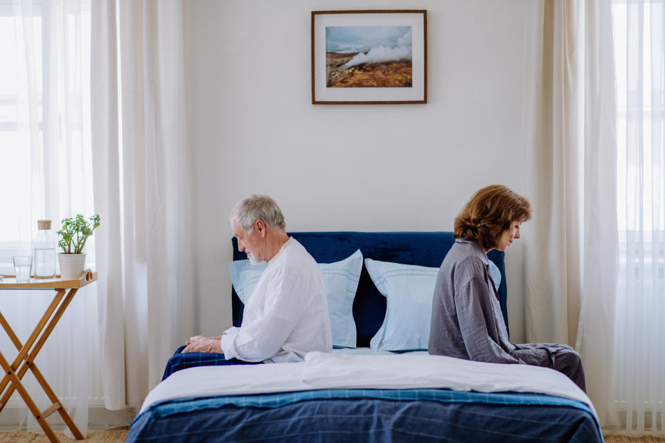 An older couple sits on opposite ends of the bed, looking away from each other, in a well-lit bedroom. A framed landscape photo hangs above the bed