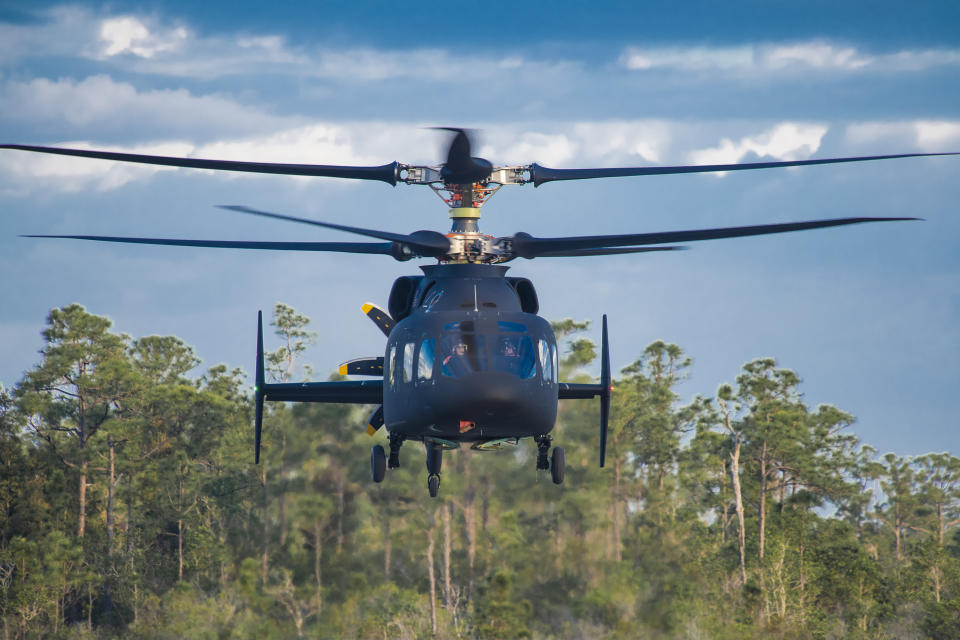 The Defiant hovers over an airfield with pine trees in the background.