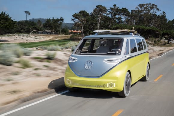 The Volkswagen I.D. Buzz concept vehicle, a silver and yellow minivan with styling inspired by the 1960s-era VW Microbus, is shown on a beach-side road in California. Two surfboards are on the vehicle's roof.