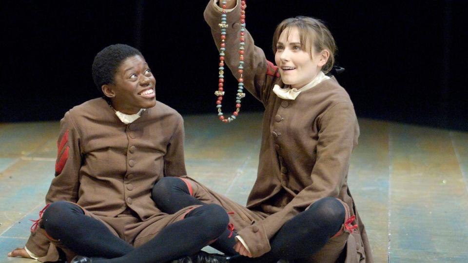 Two actors sitting on stage in 18th century costumes holding up a string of beads