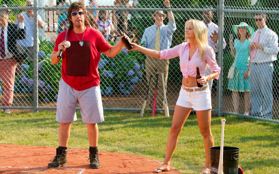Adam Sandler holds a baseball bat next to a woman in short shorts