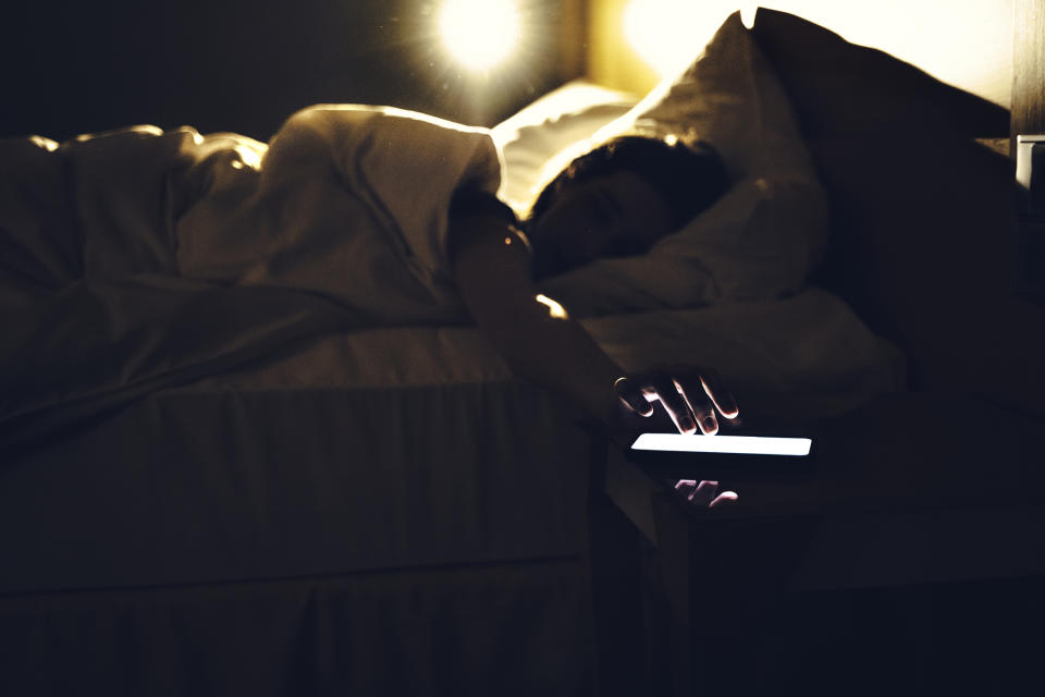 Person in bed reaching for a smartphone with illuminated screen, suggesting checking phone first thing upon waking