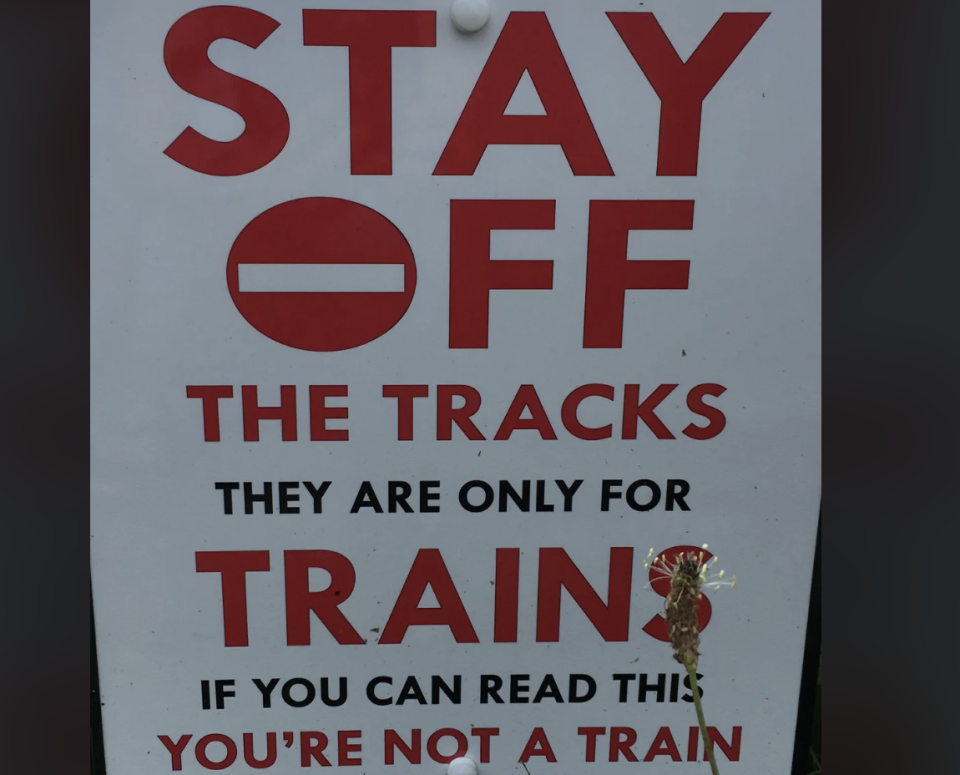 A sign with text "STAY OFF THE TRACKS THEY ARE ONLY FOR TRAINS IF YOU CAN READ THIS YOU'RE NOT A TRAIN"