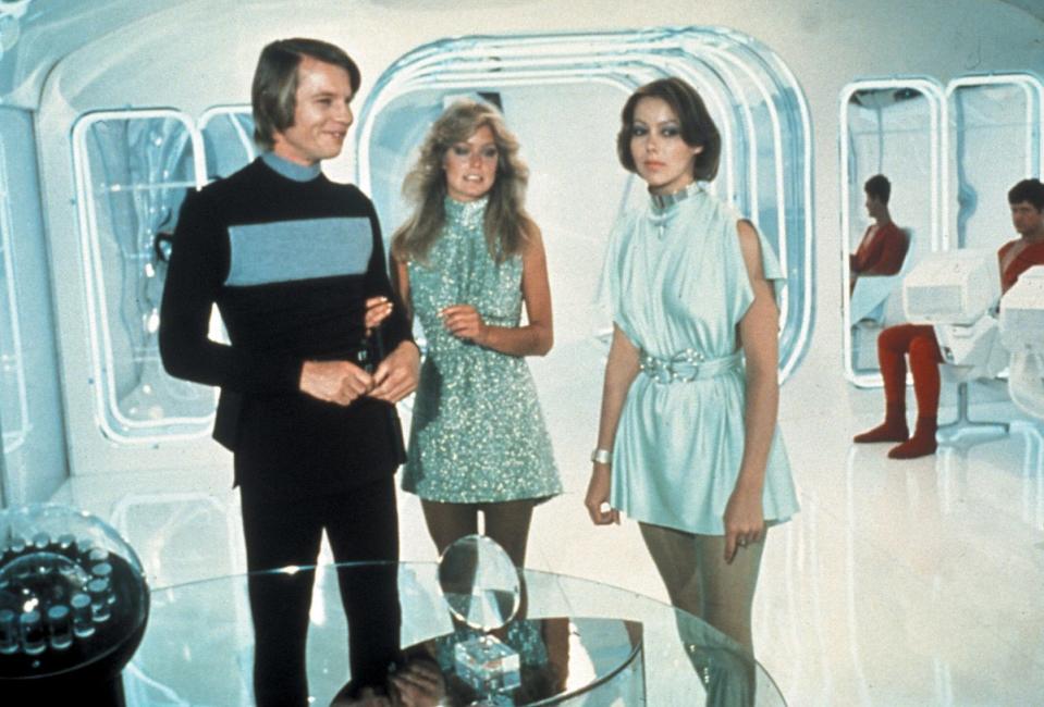 A still from Logan's Run shows Micheal York, Farrah Fawcett, and Julie Aguitter standing in skimpy silver outfits on a futuristic set