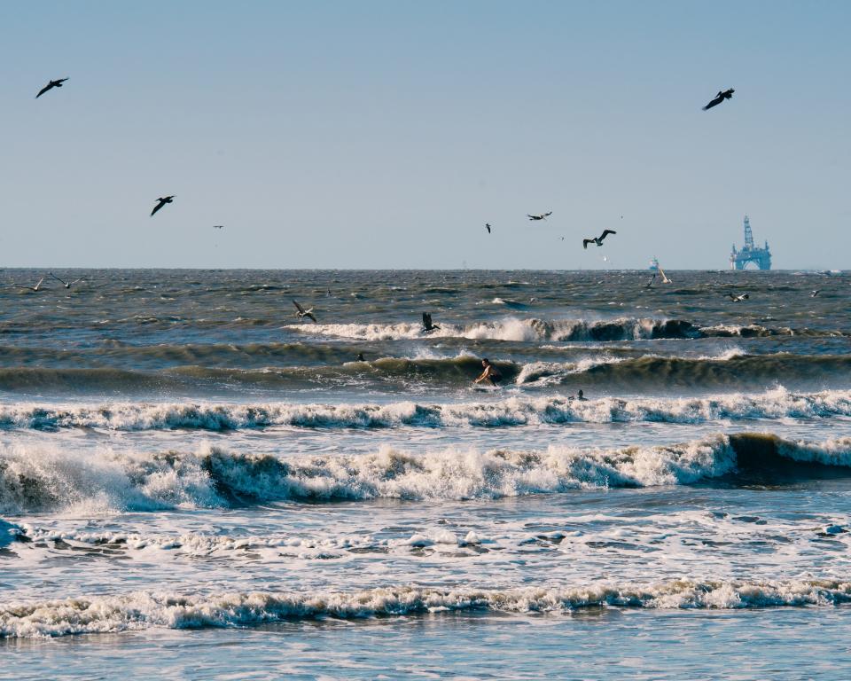 Pelicans fly overhead as a surfer finds a ride between the white wash and an oil rig looms in the background