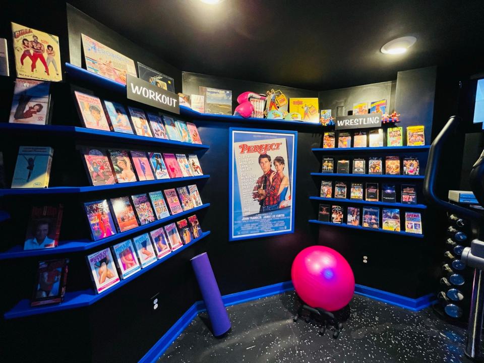 A home gym with black walls and flooring, complete with shelves of VHS tapes and workout equipment.