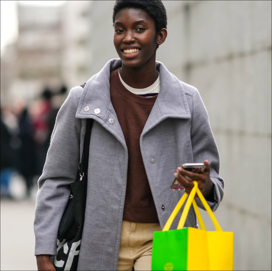  A model wears a white and navy blue striped print pattern t-shirt, a dark brown sweater, a pale gray long buttoned coat, beige large suit pants, a yellow and green shopping bag, outside Chloe, during Paris Fashion Week - Womenswear F/W 2022-2023, on March 03, 2022 in Paris, France. 