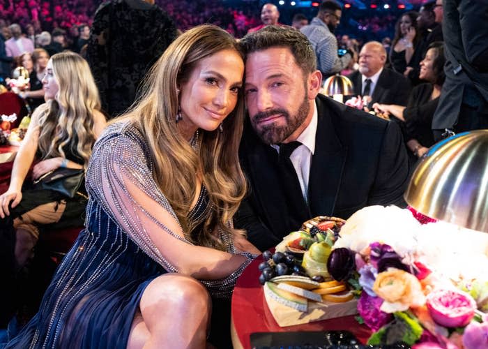 Jennifer Lopez and Ben Affleck seated at a table during an event, smiling at the camera; a variety of food items and flowers are on the table