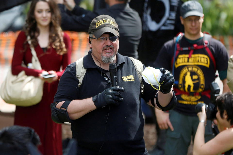 Stewart Rhodes, wearing a black baseball cap saying Oath Keepers and black gloves, at the microphone, surrounded by supporters and with a photographer taking his picture in front of him. 