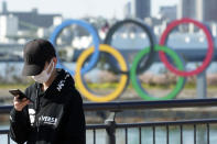 In this March 3, 2020, photo, a tourist wearing a protective mask takes a photo with the Olympic rings in the background, at Tokyo's Odaiba district in Tokyo. Japan's Olympic minister has suggested in Parliament that the Tokyo Olympics might be pushed back a few months from it July 24 opening. The games are under threat from a spreading virus from China that has reached the pandemic stage. But the so-called “Home City Contract”signed by the International Olympic Committee and Japanese officials gives the IOC wide latitude in terminating the Olympics. (AP Photo/Eugene Hoshiko)