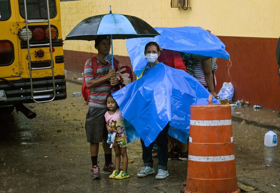 A family stands in the rain under umbrellas and tarps, behind a school bus