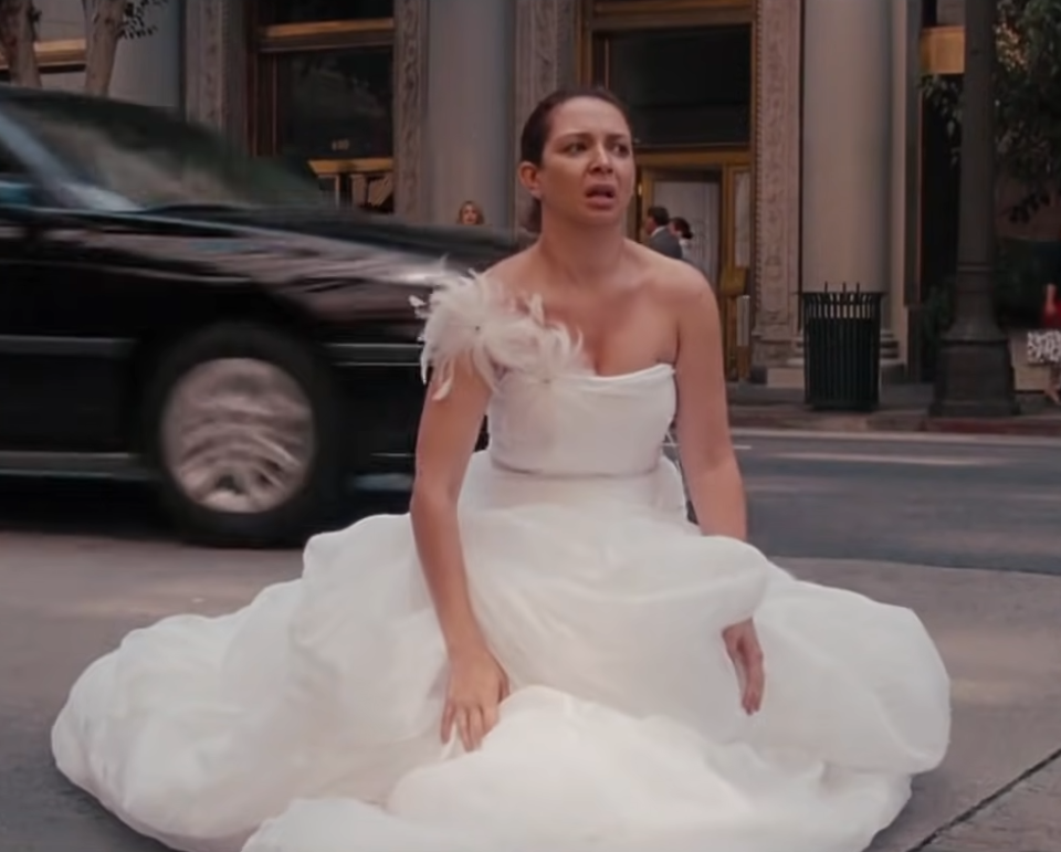 Maya Rudolph, in a white wedding dress with a feathered detail on the shoulder, sits in distress on a city street as a car passes by