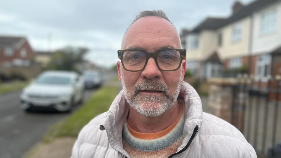 Andrew Jefferies wearing a white puffer jacket, wearing thick-rimmed glasses, standing in a residential street