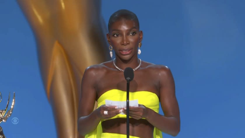 A woman in a bright yellow dress holding a piece of paper and speaking into a microphone