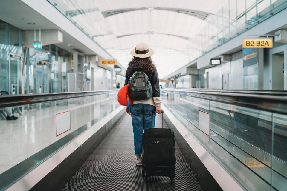 Woman wearing hat and carrying backpack and suitcase down moving walkway at airport.