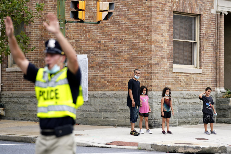 In this Sept. 9, 2020, photo pedestrians look on before crossing an intersection as a cadet in the Baltimore Police Academy directs traffic during an on the field class session in Baltimore. (AP Photo/Julio Cortez)