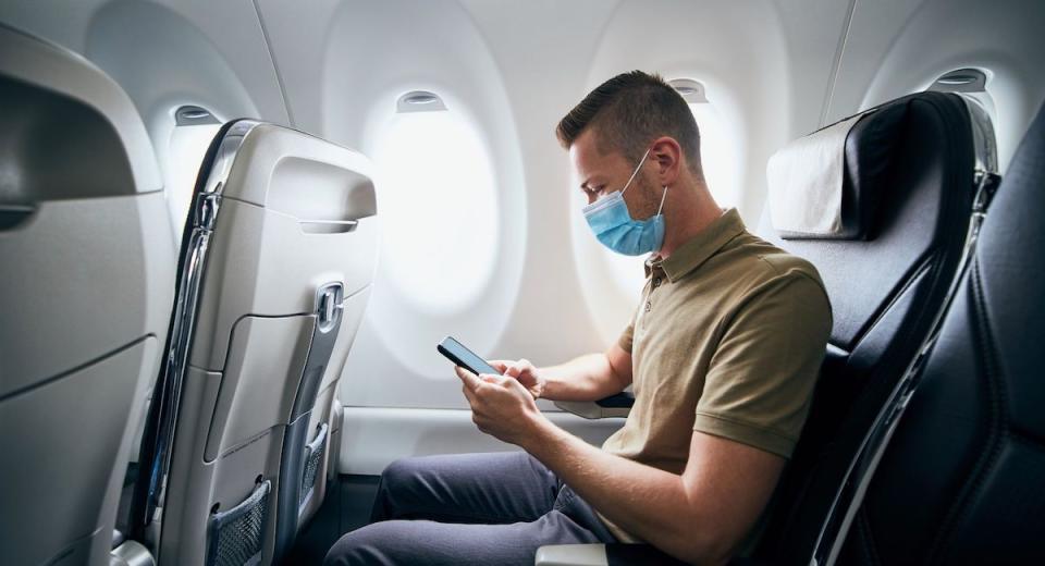 Man sitting on a window seat on a flight with a mask on and looking at a mobile phone