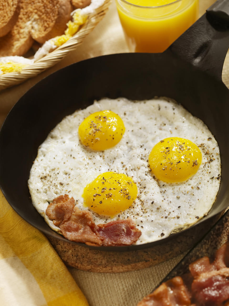 Three sunny-side-up eggs and bacon in a skillet, with a glass of orange juice and bread in the background