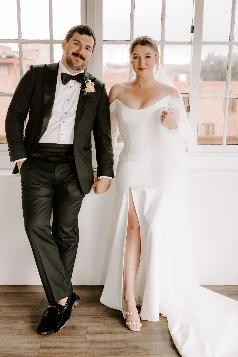 A bride and groom hold hands in front of windows.