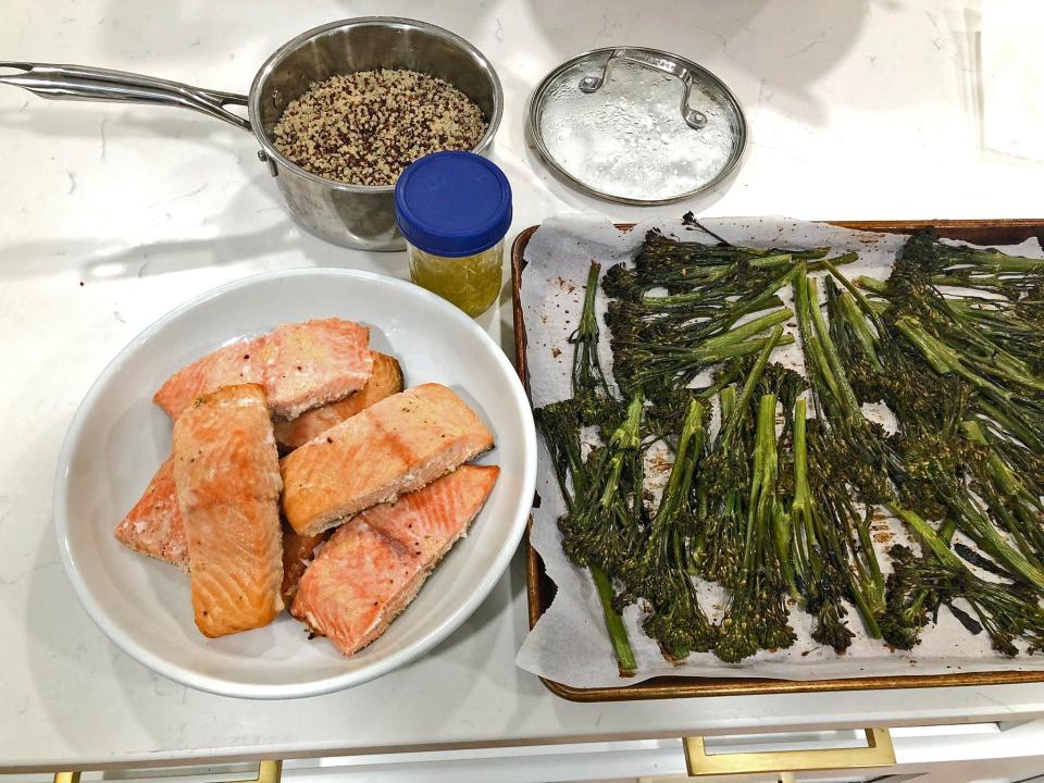 A bowl of salmon, Broccolini on a baking sheet covered in parchment paper, quinoa in a pot with the lid next to it, and a container of vinaigrette on a white countertop.