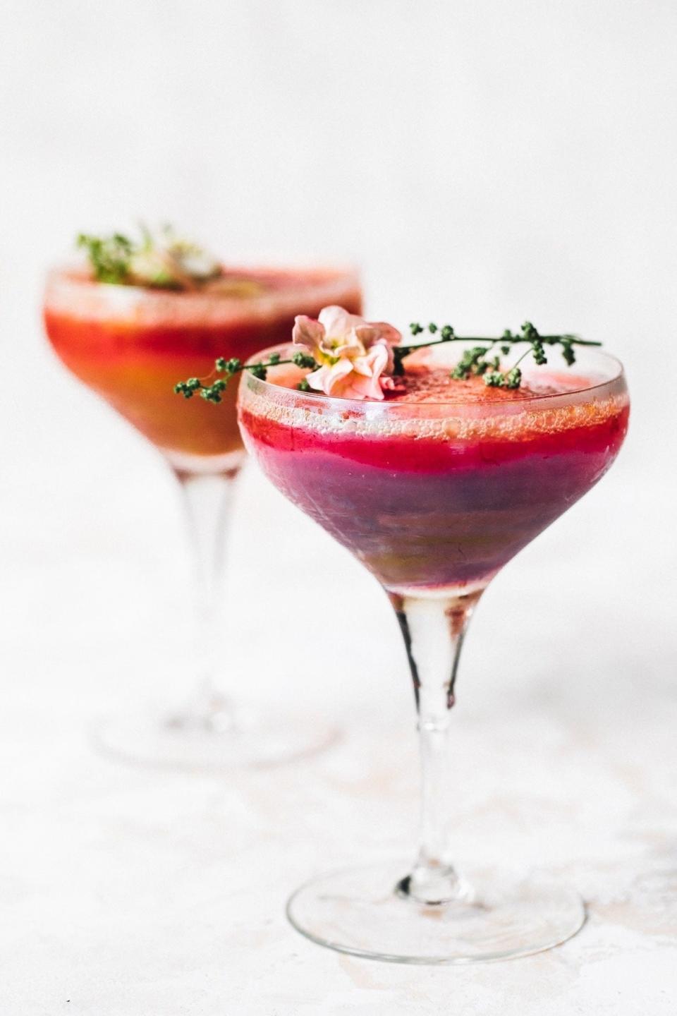 Two elegant cocktail glasses filled with layered drinks and garnished with pink flowers and sprigs of herbs