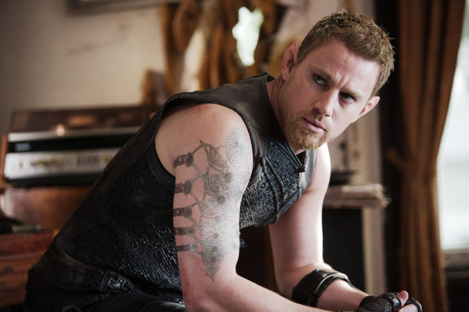 Channing Tatum is seated in a room, wearing a sleeveless, textured top, displaying a tattoo on his right arm. He appears to be in mid-conversation or thought