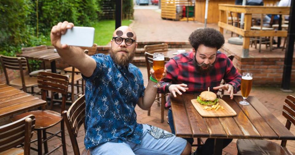 A stock image showing two men posing for a selfie while showing off food and beer